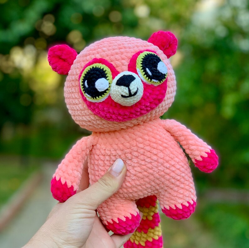 Op de afbeelding: Een roze gehaakt knuffeldier met grote zwarte ogen en een gele en roze staart. Het dier heeft een roze neus en een witte mond.