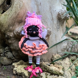 Puede incluir: Una mu&ntilde;eca de crochet con piel negra, cabello rosa y morado, un vestido rosa y melocot&oacute;n y zapatos rosas. La mu&ntilde;eca est&aacute; de pie sobre una roca con un tronco de &aacute;rbol detr&aacute;s.