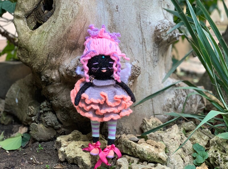 Puede incluir: Una mu&ntilde;eca de crochet con piel negra, cabello rosa y morado, un vestido rosa y melocot&oacute;n y zapatos rosas. La mu&ntilde;eca est&aacute; de pie sobre una roca con un tronco de &aacute;rbol detr&aacute;s.