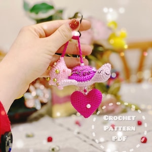 Peut inclure: Un porte-clés en forme de tortue crocheté rose et violet avec un charme en forme de cœur rose. La tortue a une carapace perlée et un visage souriant. Le porte-clés est attaché à un anneau argenté.
