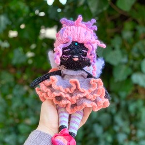 Puede incluir: Una mu&ntilde;eca de crochet con pelo rosa y morado, que lleva un vestido rosa y morado con una falda con volantes y medias a rayas. La mu&ntilde;eca tiene ojos negros y una cara negra.
