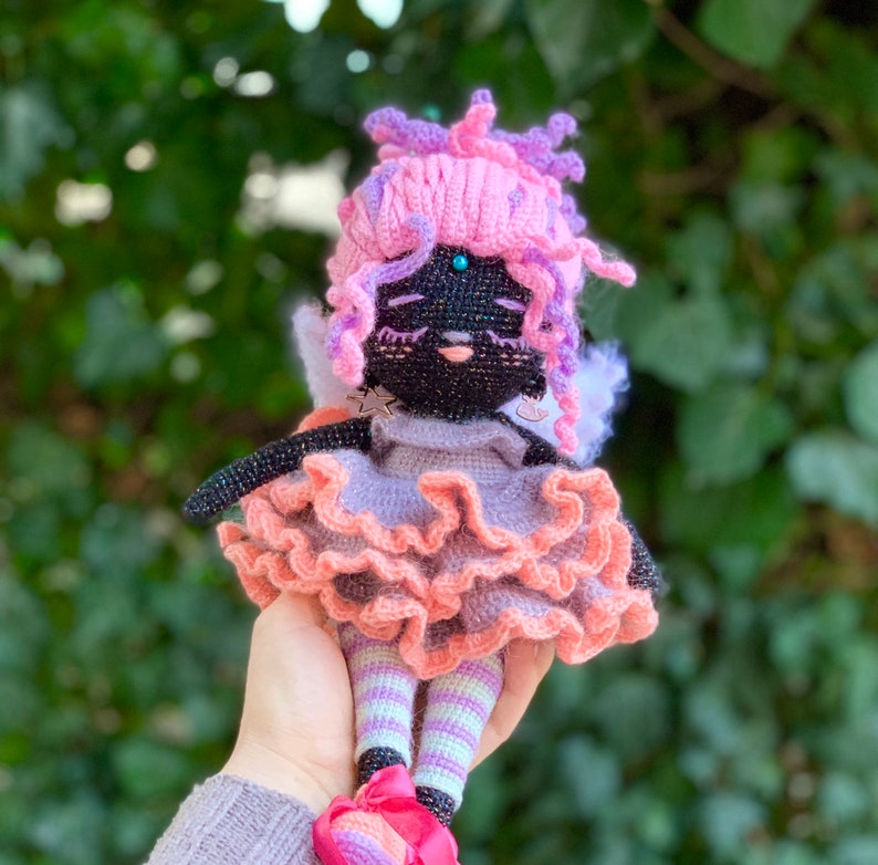 Puede incluir: Una mu&ntilde;eca de crochet con pelo rosa y morado, que lleva un vestido rosa y morado con una falda con volantes y medias a rayas. La mu&ntilde;eca tiene ojos negros y una cara negra.