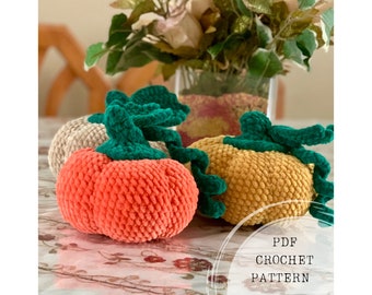 May include: Three crocheted pumpkins in orange, beige, and yellow with green stems. The pumpkins are sitting on a table with a floral arrangement in the background. PDF CROCHET PATTERN is written on a white circle in the bottom right corner of the image.