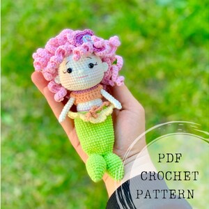 May include: Crochet pattern for a mermaid doll with pink hair, a peach-colored top, and a green tail. The doll is holding a purple flower in her hair. PDF Crochet Pattern.