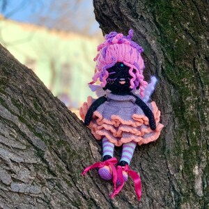 Puede incluir: Una mu&ntilde;eca de crochet con pelo rosa, cara negra y vestido morado y rosa. La mu&ntilde;eca est&aacute; sentada en un tronco de &aacute;rbol.