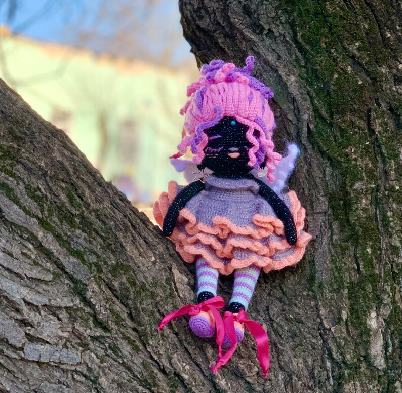 Puede incluir: Una mu&ntilde;eca de crochet con pelo rosa, cara negra y vestido morado y rosa. La mu&ntilde;eca est&aacute; sentada en un tronco de &aacute;rbol.