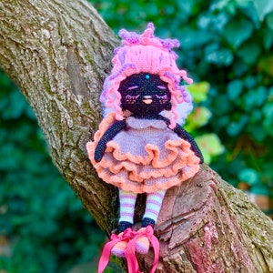 Puede incluir: Una mu&ntilde;eca de crochet con piel negra, cabello rosa y un vestido rosa y melocot&oacute;n. La mu&ntilde;eca lleva zapatos rosas y est&aacute; sentada en una rama de &aacute;rbol.