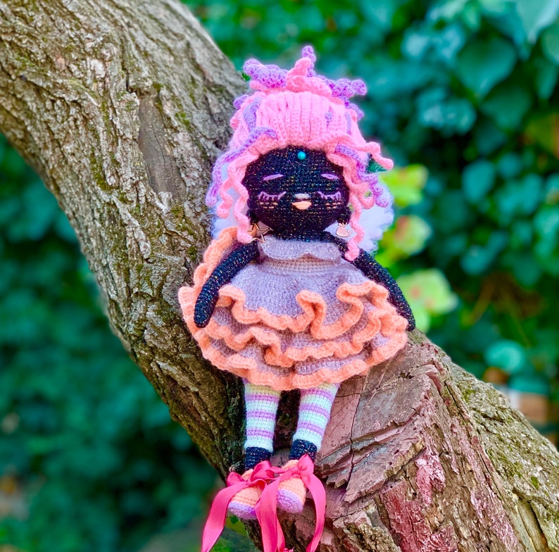 Puede incluir: Una mu&ntilde;eca de crochet con piel negra, cabello rosa y un vestido rosa y melocot&oacute;n. La mu&ntilde;eca lleva zapatos rosas y est&aacute; sentada en una rama de &aacute;rbol.