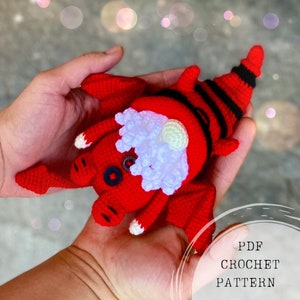 May include: A red crocheted dragon with black stripes, white yarn for the beard, and black buttons for eyes. The dragon is being held in a person's hands. The text "PDF CROCHET PATTERN" is visible in the image.