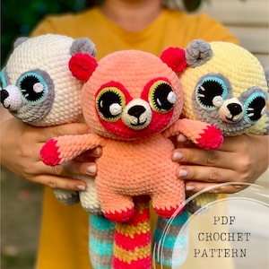 Op de afbeelding: Drie gehaakte knuffels in de vorm van wasberen. De wasberen zijn wit, roze en geel met zwarte ogen en grijze neuzen. De roze wasbeer heeft een gestreepte staart met roze, gele en blauwe strepen. De gehaakte dieren worden vastgehouden door de handen van een persoon. PDF CROCHET PATTERN staat onderaan de afbeelding.