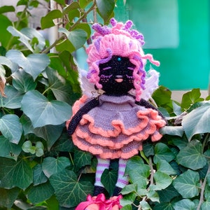 Puede incluir: Una mu&ntilde;eca de crochet con pelo rosa y morado, un vestido gris con volantes rosas y alas blancas. La mu&ntilde;eca tiene ojos negros y una cara negra.