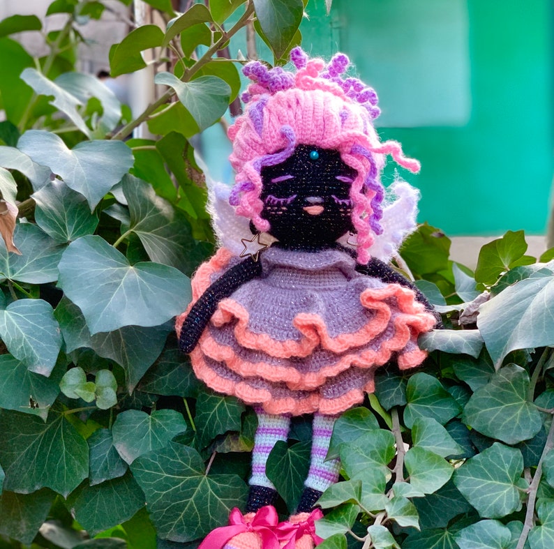 Puede incluir: Una mu&ntilde;eca de crochet con pelo rosa y morado, un vestido gris con volantes rosas y alas blancas. La mu&ntilde;eca tiene ojos negros y una cara negra.