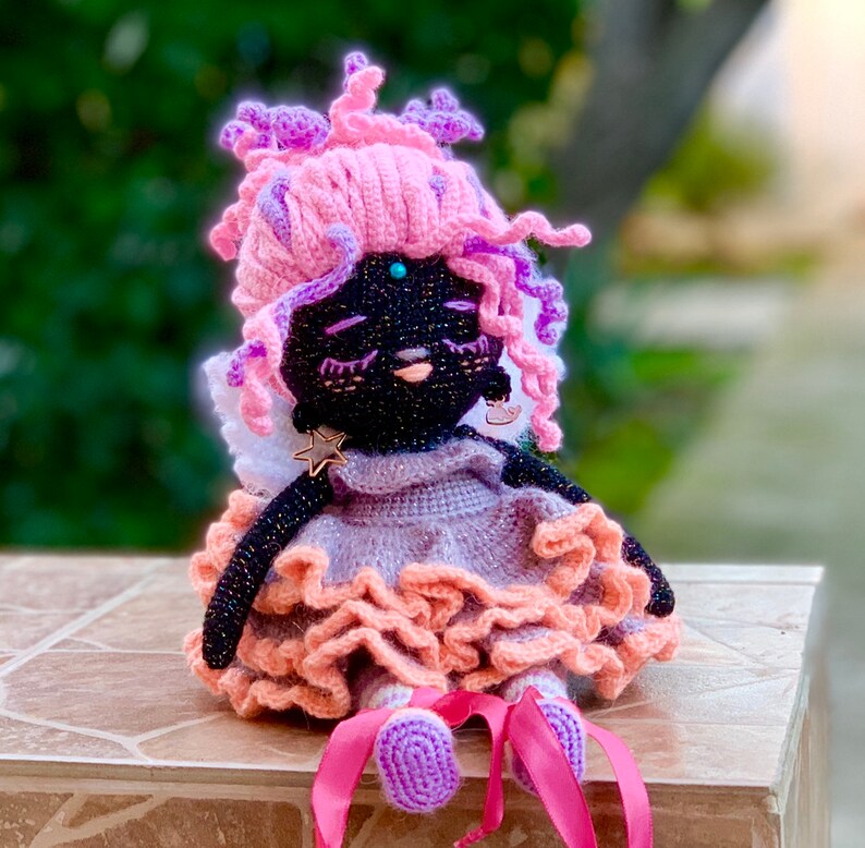 Puede incluir: Una mu&ntilde;eca de crochet con cuerpo y cara de hilo negro, pelo de hilo rosa y un vestido morado y rosa con falda de volantes. La mu&ntilde;eca lleva un amuleto en forma de estrella alrededor del cuello y tiene una gema azul claro en la frente.
