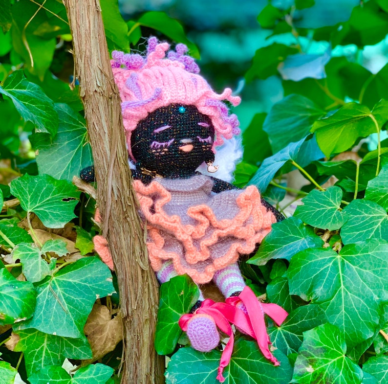 Puede incluir: Una mu&ntilde;eca de crochet con pelo rosa y morado, que lleva un vestido rosa con volantes y un cuello de encaje blanco. La mu&ntilde;eca tiene ojos negros y una nariz negra, y est&aacute; sentada en una cama de hojas verdes.
