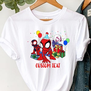Puede incluir: Camiseta blanca con una imagen de dibujos animados de Spider-Man, Spider-Gwen y otro personaje de Spider-Man sosteniendo regalos y usando sombreros de fiesta. El texto "CUSTOM TEXT" est&aacute; impreso debajo de la imagen.