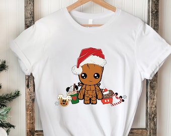 May include: A white short-sleeved t-shirt featuring a graphic of a brown tree-like character wearing a red and white Santa hat. The character is surrounded by holiday-themed items including a green cup, a cookie, and a candy cane.