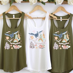 May include: Three tank tops hanging on wooden hangers. Two are olive green, and one is white. Each tank top features a colorful graphic design with a dragon, castle, and other whimsical elements. A floral arrangement of white roses hangs above.