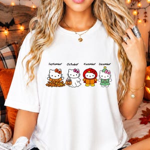 Puede incluir: Una camiseta blanca con un gr&aacute;fico de cuatro personajes de Hello Kitty etiquetados como septiembre, octubre, noviembre y diciembre. Los personajes visten atuendos de temporada que incluyen hojas de oto&ntilde;o, un disfraz de fantasma y un &aacute;rbol.