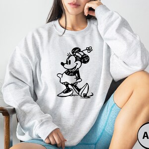 Puede incluir: Una sudadera gris claro con una ilustraci&oacute;n en blanco y negro de Minnie Mouse. La sudadera tiene cuello redondo y mangas largas. El texto "Ash" es visible en la esquina inferior derecha de la imagen.