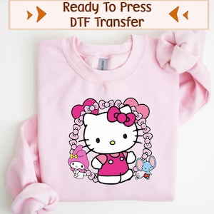 May include: A light pink sweatshirt featuring a graphic of Hello Kitty in a pink dress and bow, surrounded by a heart-shaped frame. The design includes other cartoon characters. The text "Ready To Press DTF Transfer" is at the top.