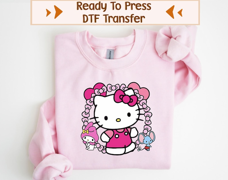 K&ouml;nnte beinhalten: Hellrosa Sweatshirt mit einem Aufdruck von Hello Kitty in einem rosa Kleid und Schleife, umgeben von einem herzf&ouml;rmigen Rahmen. Das Design beinhaltet weitere Zeichentrickfiguren. Der Text "Ready To Press DTF Transfer" befindet sich oben.