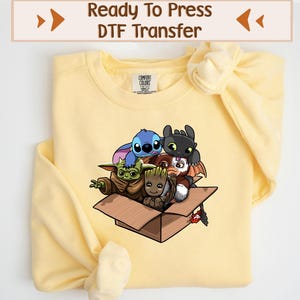 May include: A light yellow sweatshirt featuring a cartoon graphic of several animated characters inside a cardboard box. The characters include Stitch, Baby Yoda, Groot, and Toothless. The text "Ready To Press DTF Transfer" is at the top.