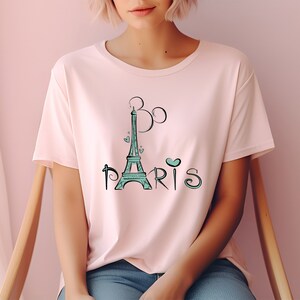 Op de afbeelding: Een lichtroze T-shirt met een turquoise Eiffeltoren-ontwerp en het woord "Paris". Het ontwerp bevat hartjes en een gestileerde "B" boven de toren. Het shirt heeft korte mouwen en een ronde hals.