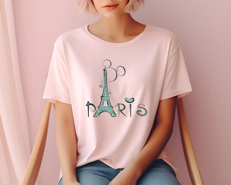 Op de afbeelding: Een lichtroze T-shirt met een turquoise Eiffeltoren-ontwerp en het woord "Paris". Het ontwerp bevat hartjes en een gestileerde "B" boven de toren. Het shirt heeft korte mouwen en een ronde hals.