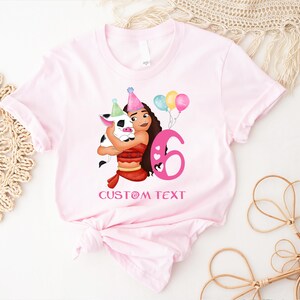 Pu&ograve; includere: Maglietta rosa con un'illustrazione di cartone animato di una ragazza con i capelli castani e un maiale che tiene palloncini e un numero 6 rosa. Il testo "CUSTOM TEXT" &egrave; stampato sotto l'illustrazione.