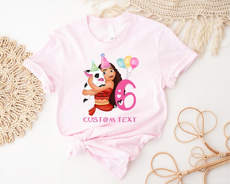 Pu&ograve; includere: Maglietta rosa con un'illustrazione di cartone animato di una ragazza con i capelli castani e un maiale che tiene palloncini e un numero 6 rosa. Il testo "CUSTOM TEXT" &egrave; stampato sotto l'illustrazione.
