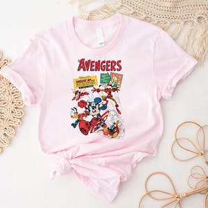 May include: A pale pink t-shirt with a comic book-style illustration of Disney characters as Avengers. The design includes Mickey Mouse, Donald Duck, and other characters in action poses, with the word "Avengers" in red.