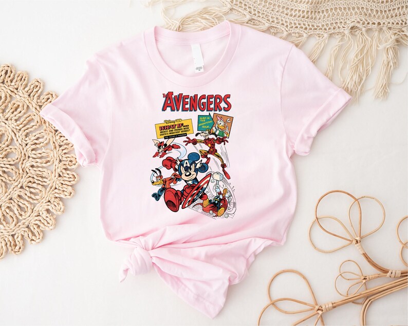 May include: A pale pink t-shirt with a comic book-style illustration of Disney characters as Avengers. The design includes Mickey Mouse, Donald Duck, and other characters in action poses, with the word "Avengers" in red.