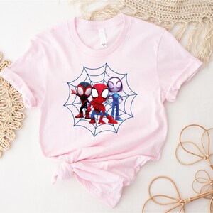 May include: Pink t-shirt with a graphic of three cartoon Spider-Man characters in a web. The characters are red, blue, and black.