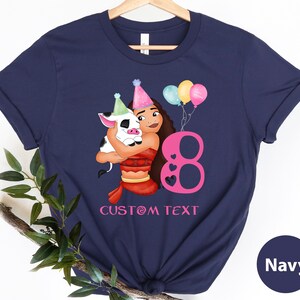 Pu&ograve; includere: Maglietta blu navy con un'illustrazione di cartone animato di una ragazza con un maiale, palloncini e il numero 8. Il testo "CUSTOM TEXT" &egrave; stampato sotto l'illustrazione.
