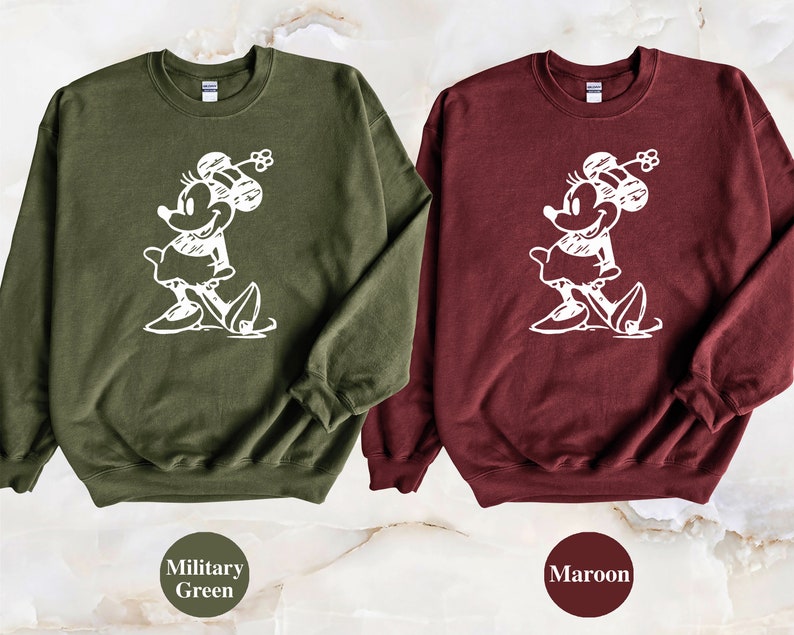 Puede incluir: Dos sudaderas con un contorno blanco de Minnie Mouse en la parte delantera. La sudadera de la izquierda es verde militar y la sudadera de la derecha es granate.