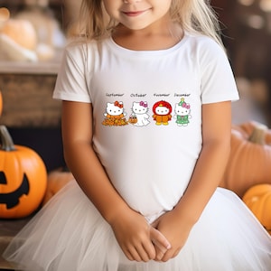 Puede incluir: Una camiseta blanca de manga corta con un estampado gr&aacute;fico de cuatro personajes de Hello Kitty que representan los meses de septiembre, octubre, noviembre y diciembre, cada uno vestido con ropa de temporada que incluye hojas de oto&ntilde;o y un disfraz.