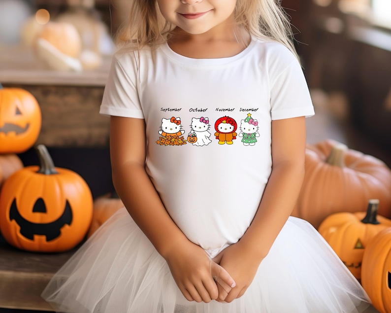 Puede incluir: Una camiseta blanca de manga corta con un estampado gr&aacute;fico de cuatro personajes de Hello Kitty que representan los meses de septiembre, octubre, noviembre y diciembre, cada uno vestido con ropa de temporada que incluye hojas de oto&ntilde;o y un disfraz.