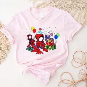 Puede incluir: Camiseta rosa con una imagen de dibujos animados de tres personajes de Spider-Man celebrando un cumplea&ntilde;os. Los personajes llevan gorros de fiesta y sostienen regalos. Hay globos y confeti en el fondo.