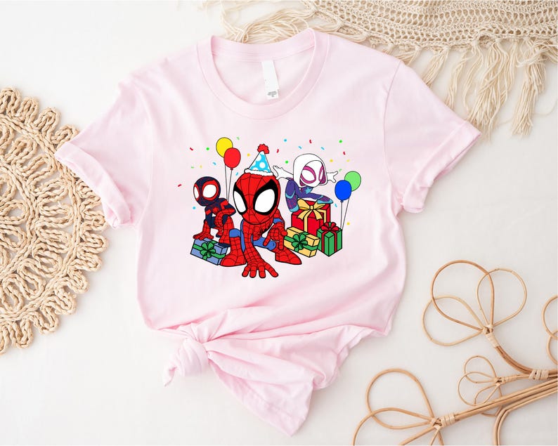 Puede incluir: Camiseta rosa con una imagen de dibujos animados de tres personajes de Spider-Man celebrando un cumplea&ntilde;os. Los personajes llevan gorros de fiesta y sostienen regalos. Hay globos y confeti en el fondo.