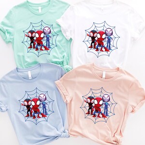 May include: Four t-shirts with a graphic print of three cartoon Spider-Man characters standing on a web. The shirts are in different colors: light blue, light green, white, and pink.