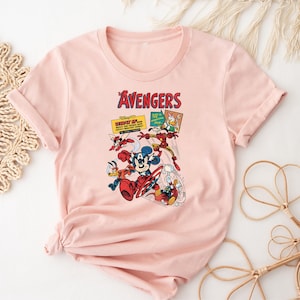 May include: A pale pink t-shirt with a graphic of Disney's Avengers characters. The design includes Mickey Mouse as Captain America, Donald Duck, and Iron Man. The word "Avengers" is printed in red above the characters.