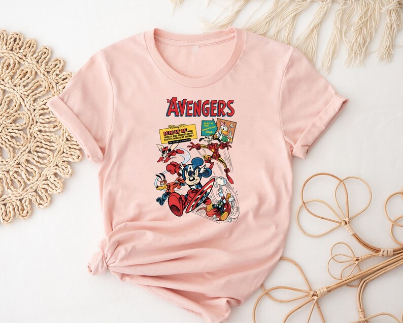 May include: A pale pink t-shirt with a graphic of Disney's Avengers characters. The design includes Mickey Mouse as Captain America, Donald Duck, and Iron Man. The word "Avengers" is printed in red above the characters.