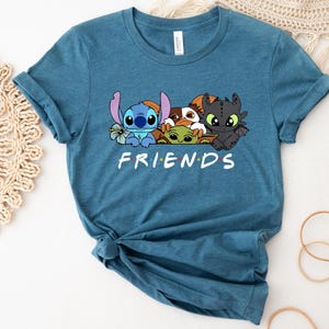May include: Teal blue t-shirt featuring a graphic of cartoon characters Stitch, Baby Yoda, Toothless, and others. The word "FRIENDS" is printed in white below the characters. The shirt is made of a soft material and has rolled-up sleeves.