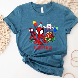 Puede incluir: Una camiseta azul turquesa con una imagen de dibujos animados de Spider-Man, Spider-Gwen y Miles Morales celebrando un cumplea&ntilde;os con regalos y globos. El texto "CUSTOM TEXT" est&aacute; impreso debajo de la imagen.
