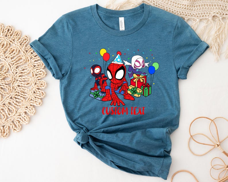 Puede incluir: Una camiseta azul turquesa con una imagen de dibujos animados de Spider-Man, Spider-Gwen y Miles Morales celebrando un cumplea&ntilde;os con regalos y globos. El texto "CUSTOM TEXT" est&aacute; impreso debajo de la imagen.