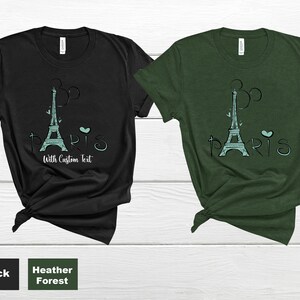 Op de afbeelding: Zwarte en heidegroene t-shirts met een turquoise Eiffeltoren-ontwerp en het woord "Paris". De tekst "With Custom Text" is ook inbegrepen. De shirts zijn gemaakt van een zacht materiaal.