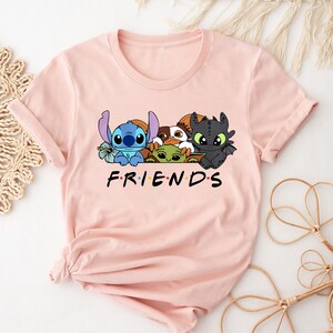 May include: A light pink t-shirt featuring a graphic of cartoon characters, including Stitch, Toothless, and others. The word "FRIENDS" is printed in black block letters below the characters. The shirt has rolled-up sleeves.