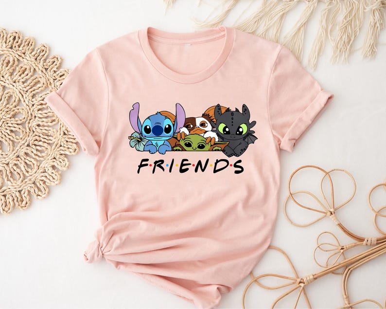 May include: A light pink t-shirt featuring a graphic of cartoon characters, including Stitch, Toothless, and others. The word "FRIENDS" is printed in black block letters below the characters. The shirt has rolled-up sleeves.