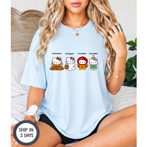 Puede incluir: Una camiseta azul claro con un estampado gr&aacute;fico de cuatro personajes de Hello Kitty etiquetados como September, October, November y December. El personaje de septiembre est&aacute; entre hojas, octubre es un fantasma, noviembre lleva una capucha roja.