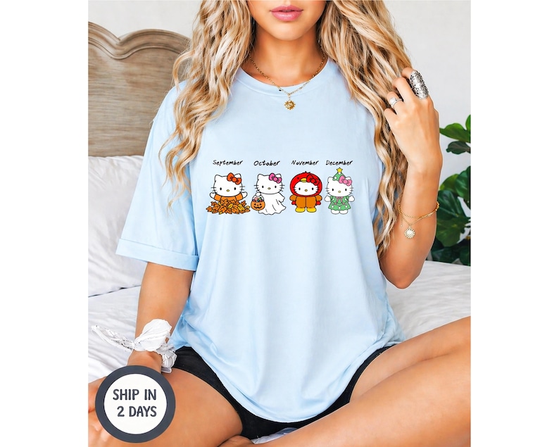 Puede incluir: Una camiseta azul claro con un estampado gr&aacute;fico de cuatro personajes de Hello Kitty etiquetados como September, October, November y December. El personaje de septiembre est&aacute; entre hojas, octubre es un fantasma, noviembre lleva una capucha roja.